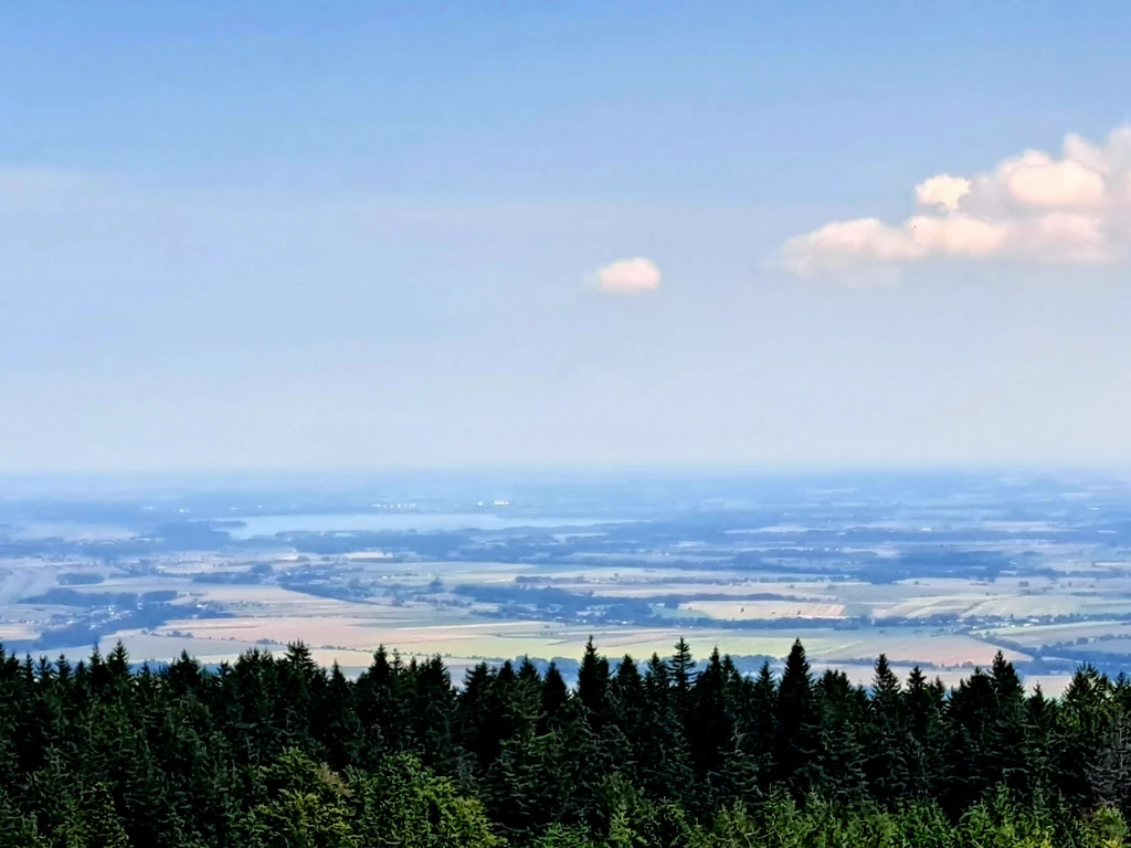 Z dolnośląskich szczytów rozpościerają się zapierające dech w piersiach widoki Panorama rozległych pól i łąk rozciągających się za zielonym, gęstym lasem świerkowym, na niebie widoczne pojedyncze chmury oraz łagodny błękit.