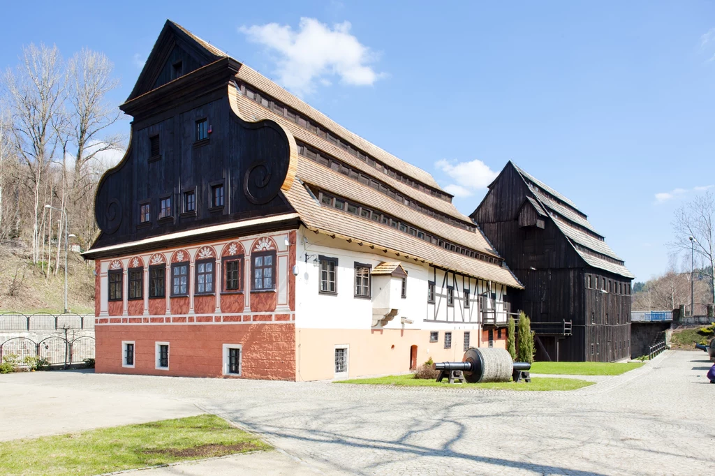Muzeum Papiernictwa w Dusznikach-Zdroju Duży historyczny budynek z charakterystyczną czarną, drewnianą fasadą i białą dolną częścią z czerwonym cokołem, stojący na otwartej przestrzeni, obok mniejszy drewniany obiekt, wokół drzewa bez liści i wybrukowane podwórze.