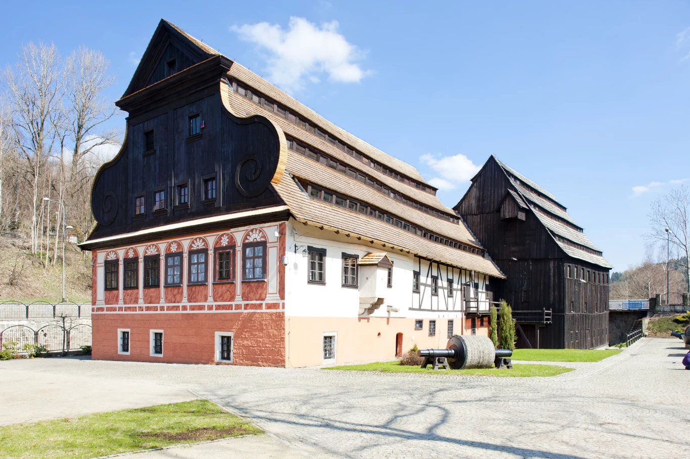 Muzeum Papiernictwa w Dusznikach-Zdroju Duży historyczny budynek z charakterystyczną czarną, drewnianą fasadą i białą dolną częścią z czerwonym cokołem, stojący na otwartej przestrzeni, obok mniejszy drewniany obiekt, wokół drzewa bez liści i wybrukowane podwórze.