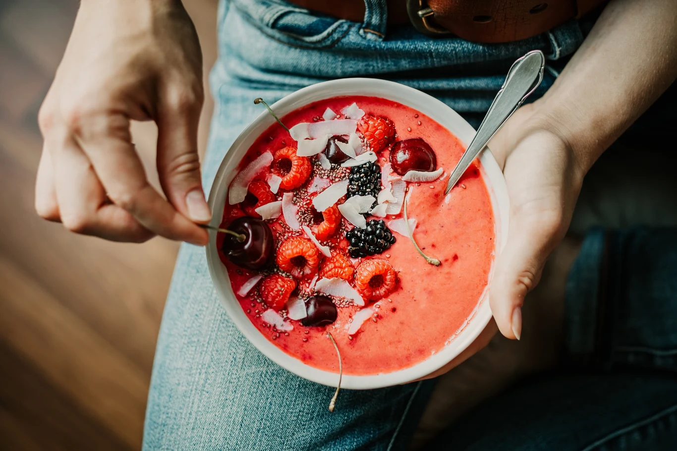 Miska gęstego smoothie w intensywnie czerwonym kolorze z dodatkiem malin, jeżyn, wiśni oraz płatków kokosowych, trzymana na kolanach osoby w dżinsach; jedna ręka trzyma miseczkę, a druga sięga po owoc.