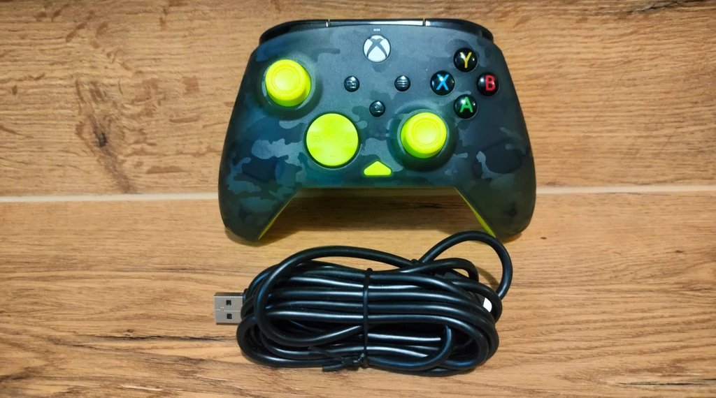 Czarny kontroler do gier z neonowo-żółtymi przyciskami oraz analogami leży na drewnianej powierzchni, poniżej widoczny zwinięty kabel USB.