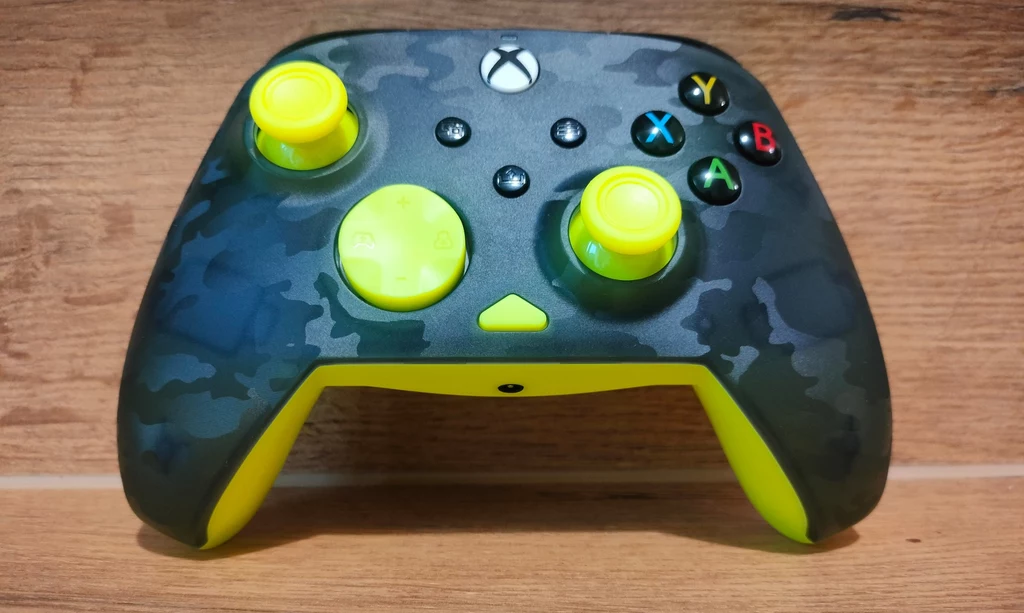 Pad do konsoli Xbox o ciemnym, moro wzorze z żółtymi elementami, leżący na drewnianej powierzchni.