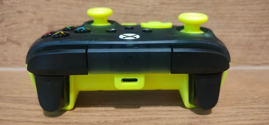Czarny kontroler do konsoli Xbox z żółtymi joystickami i detalami, ustawiony na drewnianym tle, widok od strony gniazda ładowania USB-C.