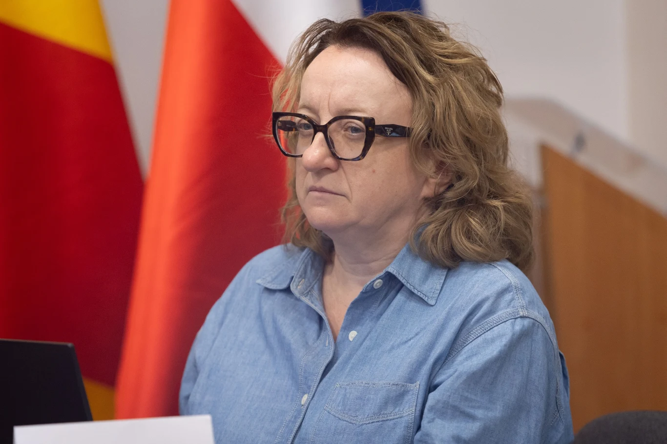 Joanna Kopczyńska odwołana z Wód Polskich Kobieta w okularach i niebieskiej koszuli siedząca przy stole na tle flag narodowych.