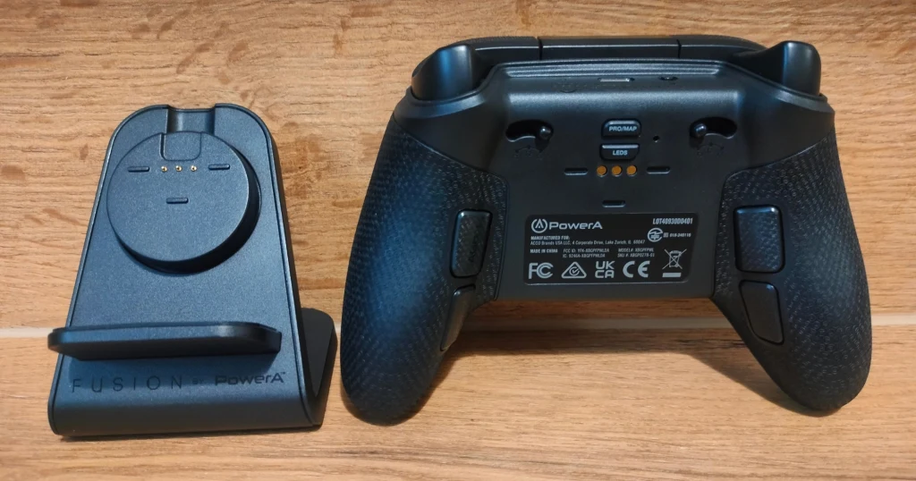 Bezprzewodowy kontroler do gier marki PowerA wraz ze stacją ładującą umieszczone są na drewnianej powierzchni, z tyłu widać teksty i oznaczenia producenta.