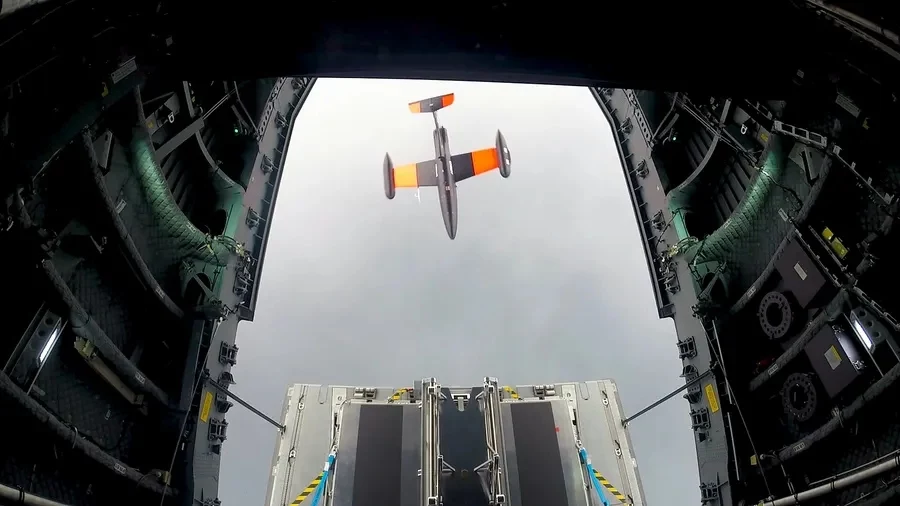 Airbus prowadził testy A400M z dronami w 2022 r. Dron z pomarańczowymi elementami unoszący się w powietrzu po wyrzuceniu go z wnętrza dużego samolotu transportowego, widok z perspektywy wnętrza samolotu na otwartą rampę i wyłaniającego się drona na tle pochmurnego nieba.