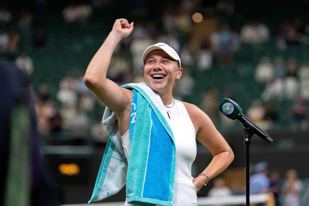 Amanda Anisimova rywalizowała z Anastazją Pawluczenkową o awans do półfinału Wimbledonu Sportowczyni w białej odzieży sportowej i czapce z daszkiem uśmiecha się szeroko, wznosząc pięść w geście triumfu, z niebieskim ręcznikiem przewieszonym przez ramię, stojąc przy mikrofonie na korcie tenisowym.