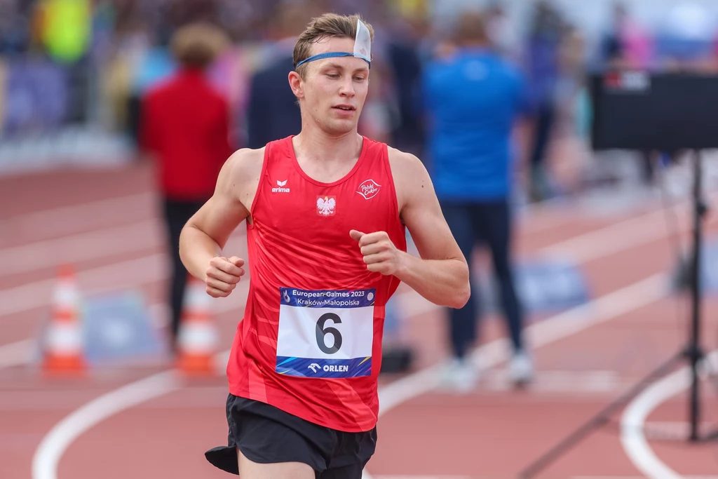 Lekkoatleta w czerwonym stroju z numerem startowym 6 biegnie po bieżni podczas zawodów sportowych, na głowie opaska chłodząca, w tle rozmazane postaci i elementy stadionu.