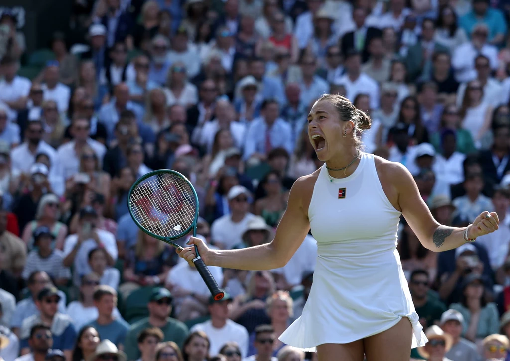 Aryna Sabalenka Zawodniczka tenisowa w białej sukience świętuje zwycięski punkt na korcie, trzymając rakietę i wykrzykując z radości, w tle widać tłum kibiców.