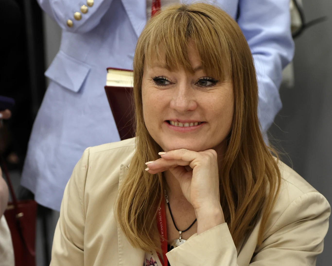 Swietłana Żurowa Kobieta o blond włosach uśmiecha się, mając rękę opartą na brodzie, ubrana w jasną marynarkę, na szyi czerwona smycz, w tle widoczne są rozmyte postacie i książka.