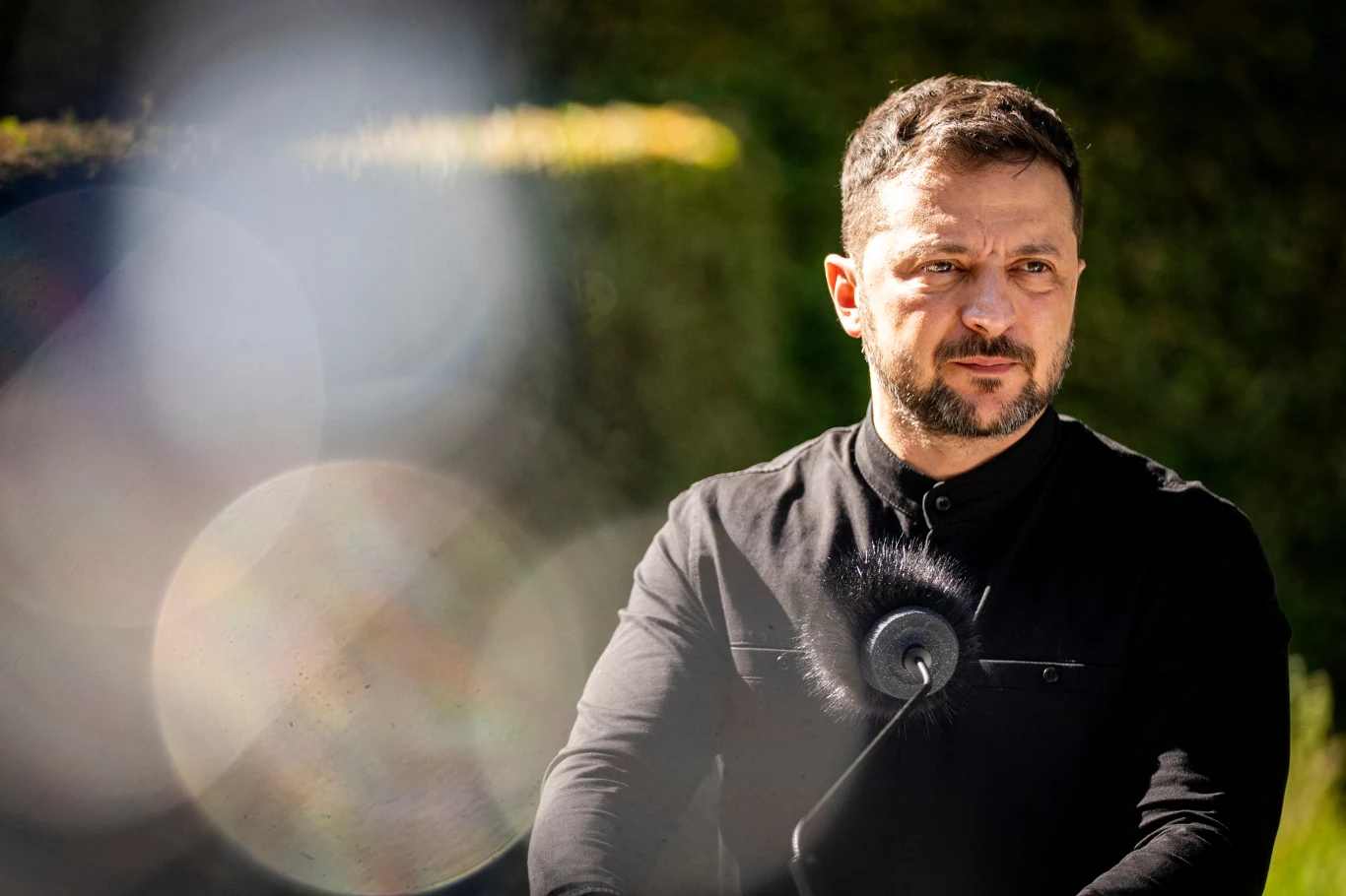 Wołodymyr Zełenski dąży do zacieśnienia więzi z USA Mężczyzna w czarnej koszuli stoi na zewnątrz, z poważnym wyrazem twarzy, tło stanowią rozmyte rośliny oraz plamy światła.