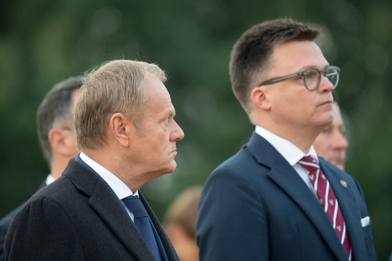Donald Tusk zabrał głos w sprawie "wyjaśnień" Szymona Hołowni Dwie dorosłe osoby w garniturach stoją obok siebie na tle rozmytego zielonego otoczenia, obie poważne i skupione, jedna w okularach, obie zwrócone w lewo.