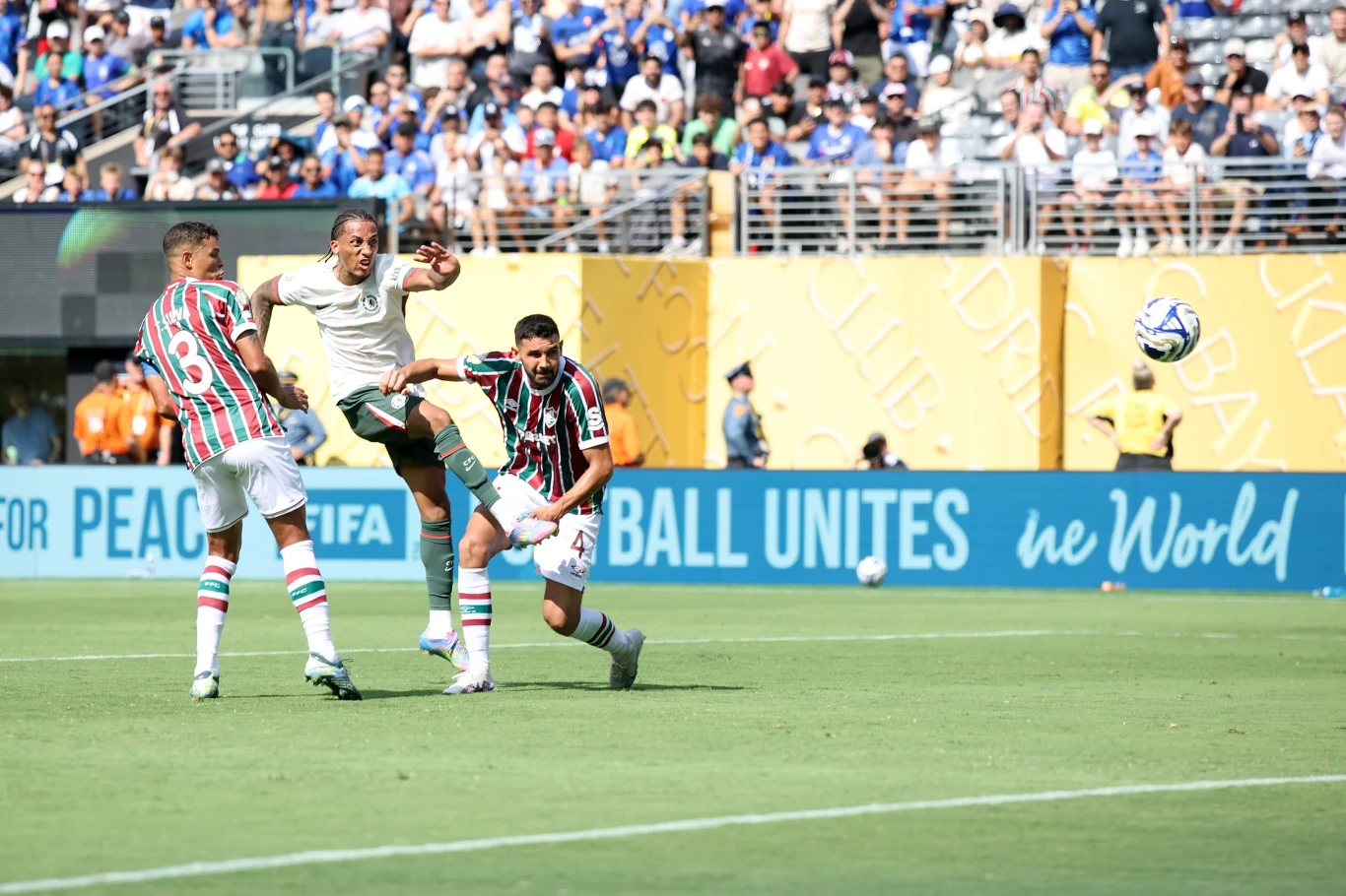 Joao Pedro strzelający gola na 2:0 Dwóch piłkarzy w pasiastych strojach próbuje zablokować zawodnika drużyny przeciwnej, który oddaje silny strzał na bramkę podczas meczu rozegranego na stadionie w słoneczny dzień. W tle widoczna licznie zgromadzona publiczność.