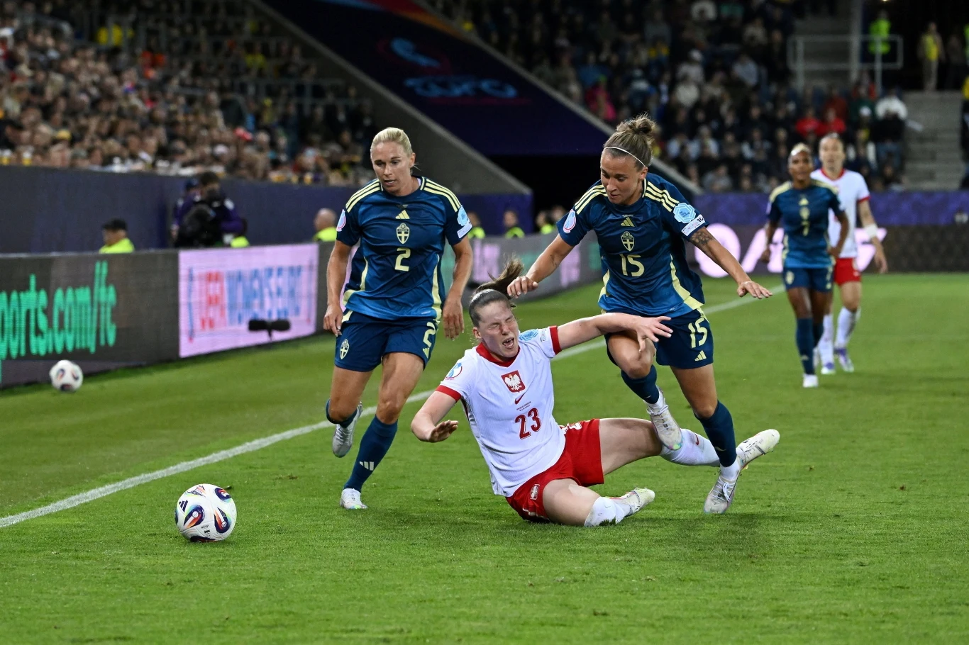 Piłkarka w biało-czerwonym stroju przewraca się na murawie boiska, naciskana przez dwie zawodniczki drużyny przeciwnej w ciemnoniebieskich strojach, w tle widoczna publiczność i fragment stadionu.