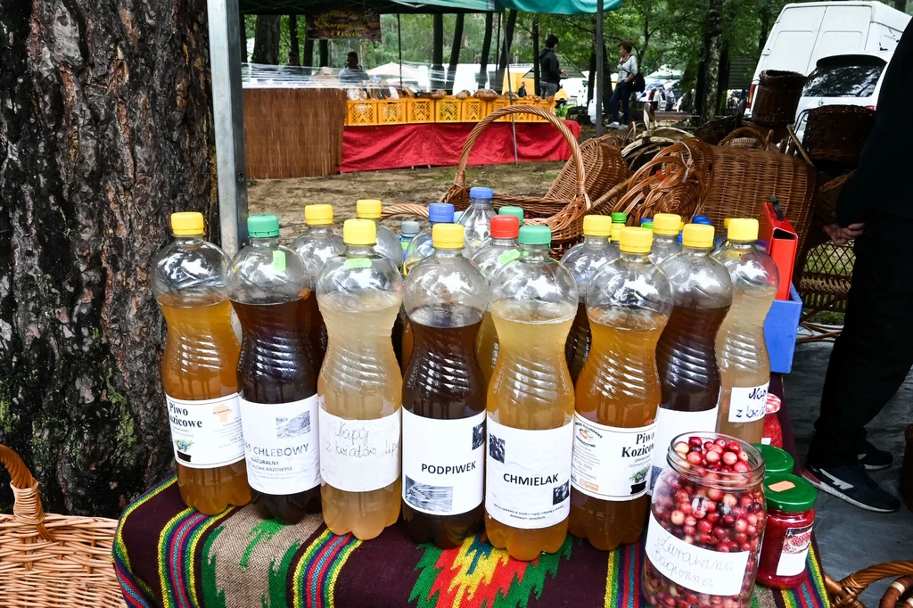 Naturalny napój - podpiwek Półtoralitrowe plastikowe butelki z domowymi napojami, takimi jak podpiwek oraz kwas chlebowy, ustawione na kolorowo zdobionym stole, w tle stoiska targowe z wiklinowymi koszami, miodami i świeżymi owocami.