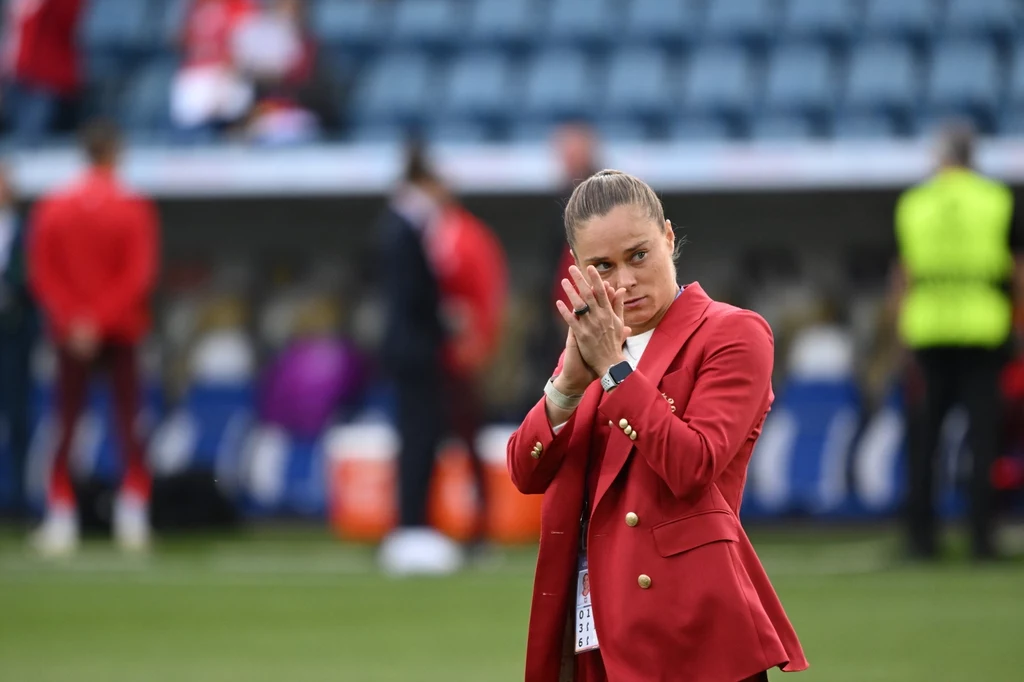 Ewa Pajor Kobieta w czerwonej marynarce stoi na murawie stadionu i klaszcze, w tle rozmazane postacie oraz trybuny z niebieskimi krzesełkami