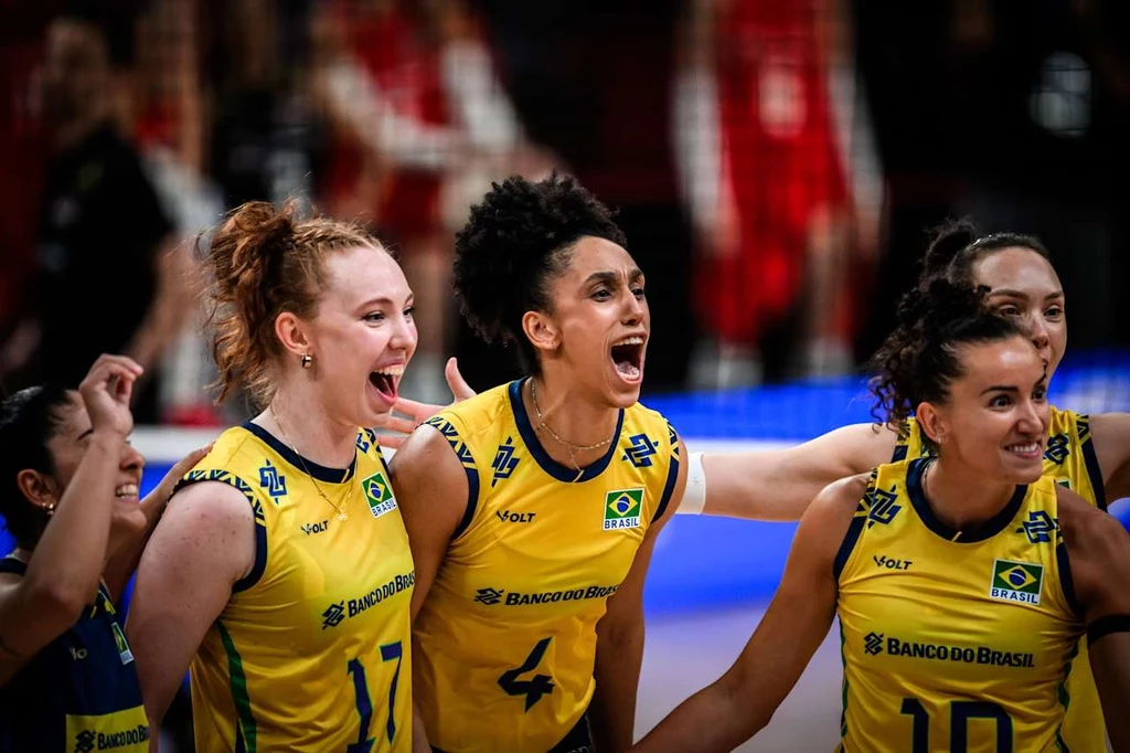 Reprezentantki Brazylii w siatkówce kobiet Trzy zawodniczki brazylijskiej drużyny siatkarskiej w żółto-niebieskich strojach świętujące punkt podczas meczu, wyrażające radość oraz zaangażowanie, z dynamicznym tłem i rozmazanymi sylwetkami rywalek w czerwonych strojach