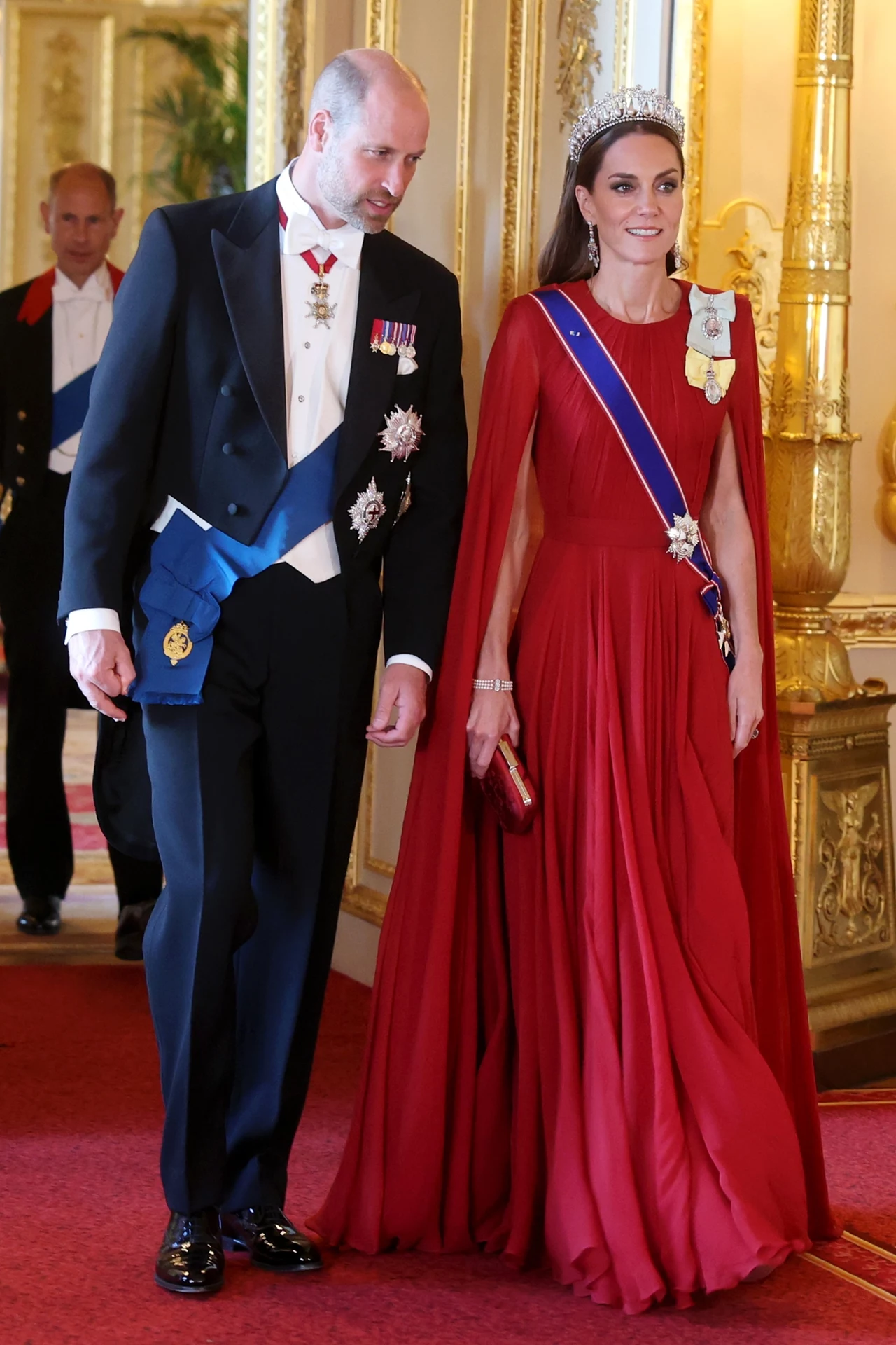 Dwie elegancko ubrane osoby idące korytarzem, mężczyzna w fraku z odznaczeniami i niebieską szarfą, kobieta w długiej czerwonej sukni z tiarą i niebieską szarfą, bogato zdobione wnętrze pałacu.