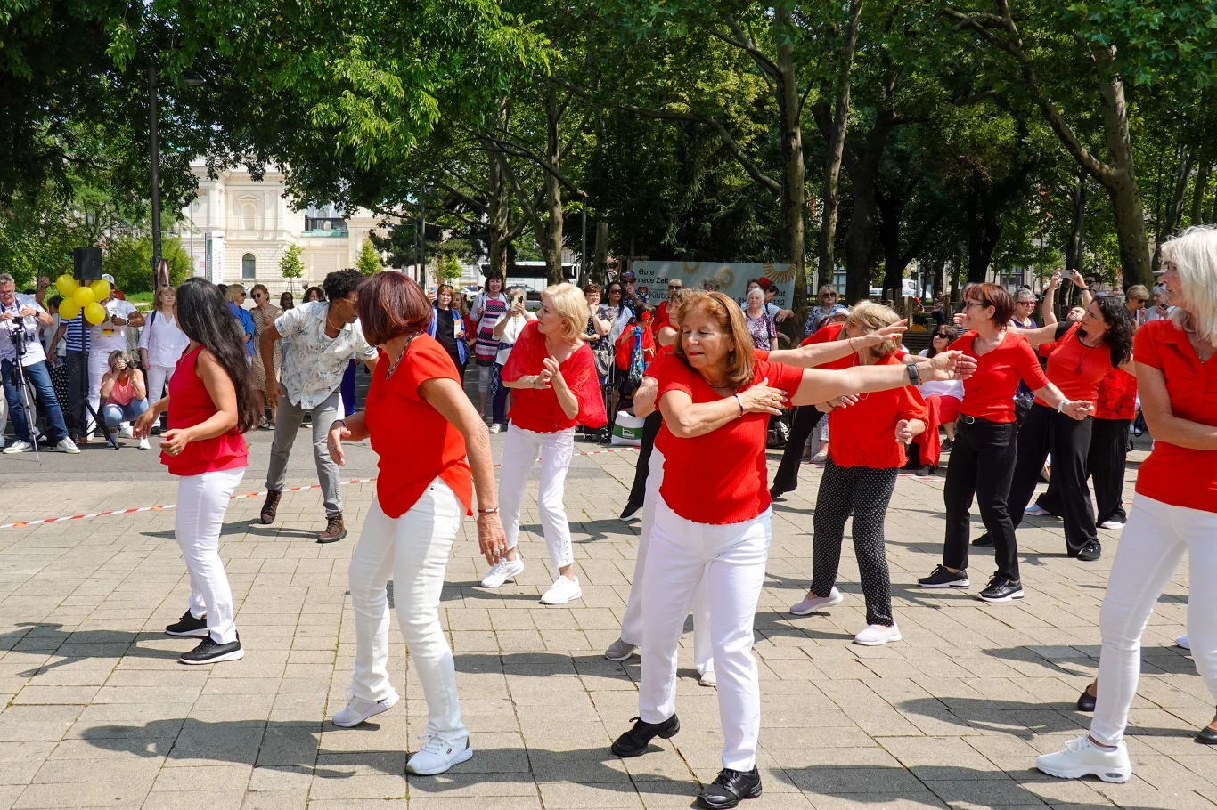 Grupa osób tańczących w rytmie na świeżym powietrzu, większość ubrana w czerwone koszulki i białe spodnie, wokół zgromadzeni widzowie, w tle drzewa i parkowe otoczenie.