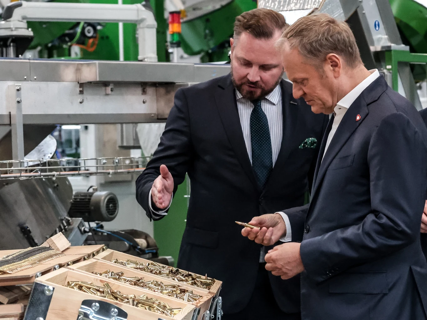 Dwaj elegancko ubrani mężczyźni stoją przy skrzynkach z amunicją, z czego jeden z nich prezentuje drugiemu nabój, wskazując na szczegóły produkcji podczas oprowadzania po nowoczesnym zakładzie przemysłowym.