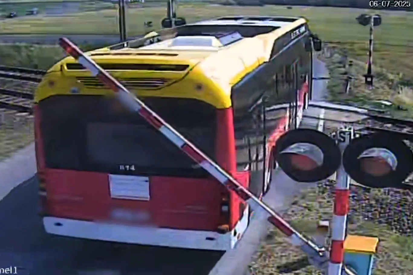 Kierowca autobusu miejskiego zignorował czerwone światło na przejeździe kolejowym. Czerwono-żółty autobus przejeżdża przez przejazd kolejowy z opuszczonym szlabanem, w tle widoczne są tory kolejowe, otoczenie stanowią pola i zieleń, scena zarejestrowana przez kamerę monitoringu.