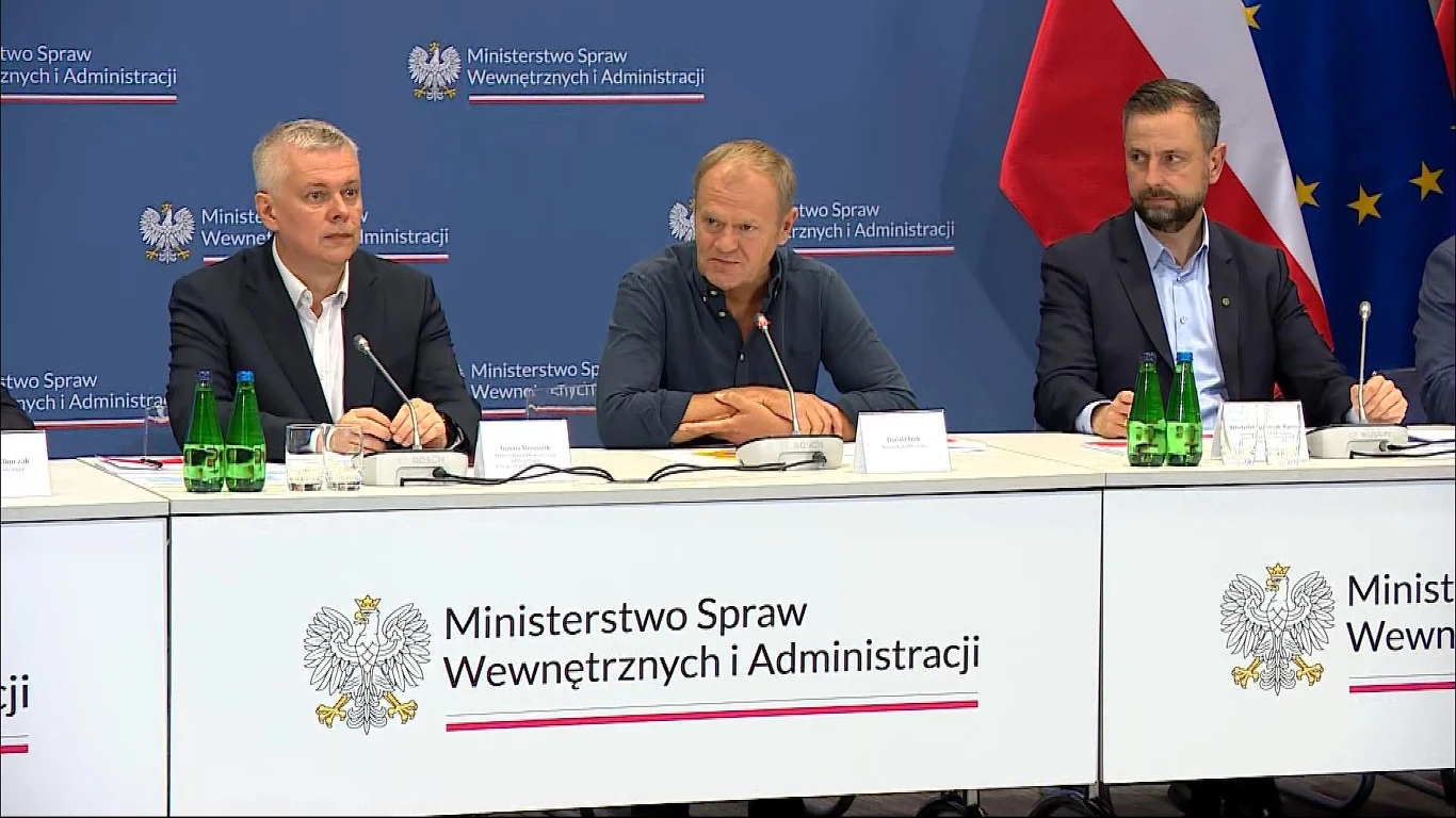 Sztab kryzysowy z udziałem premiera Donalda Tuska Trzech mężczyzn przy stole konferencyjnym podczas oficjalnej konferencji prasowej w Ministerstwie Spraw Wewnętrznych i Administracji, w tle widoczne flagi Polski oraz Unii Europejskiej.
