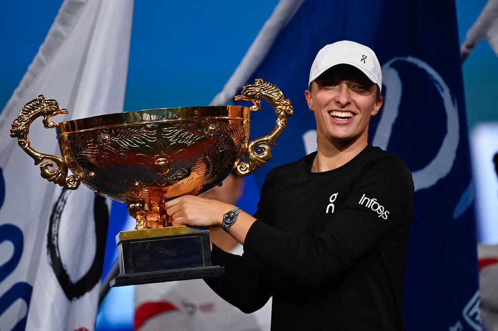 Iga Świątek z trofeum po pokonaniu Ludmiły Samsonowej w Pekinie Uśmiechnięta tenisistka w sportowej odzieży trzyma duży złoty puchar, w tle widoczne flagi oraz fragmenty innych osób, atmosfera zwycięstwa i radości
