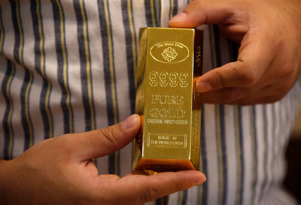 Ręce trzymają duży, błyszczący sztabek złota oznaczony napisem 9999 pure gold, na tle pasiastych ubrań.