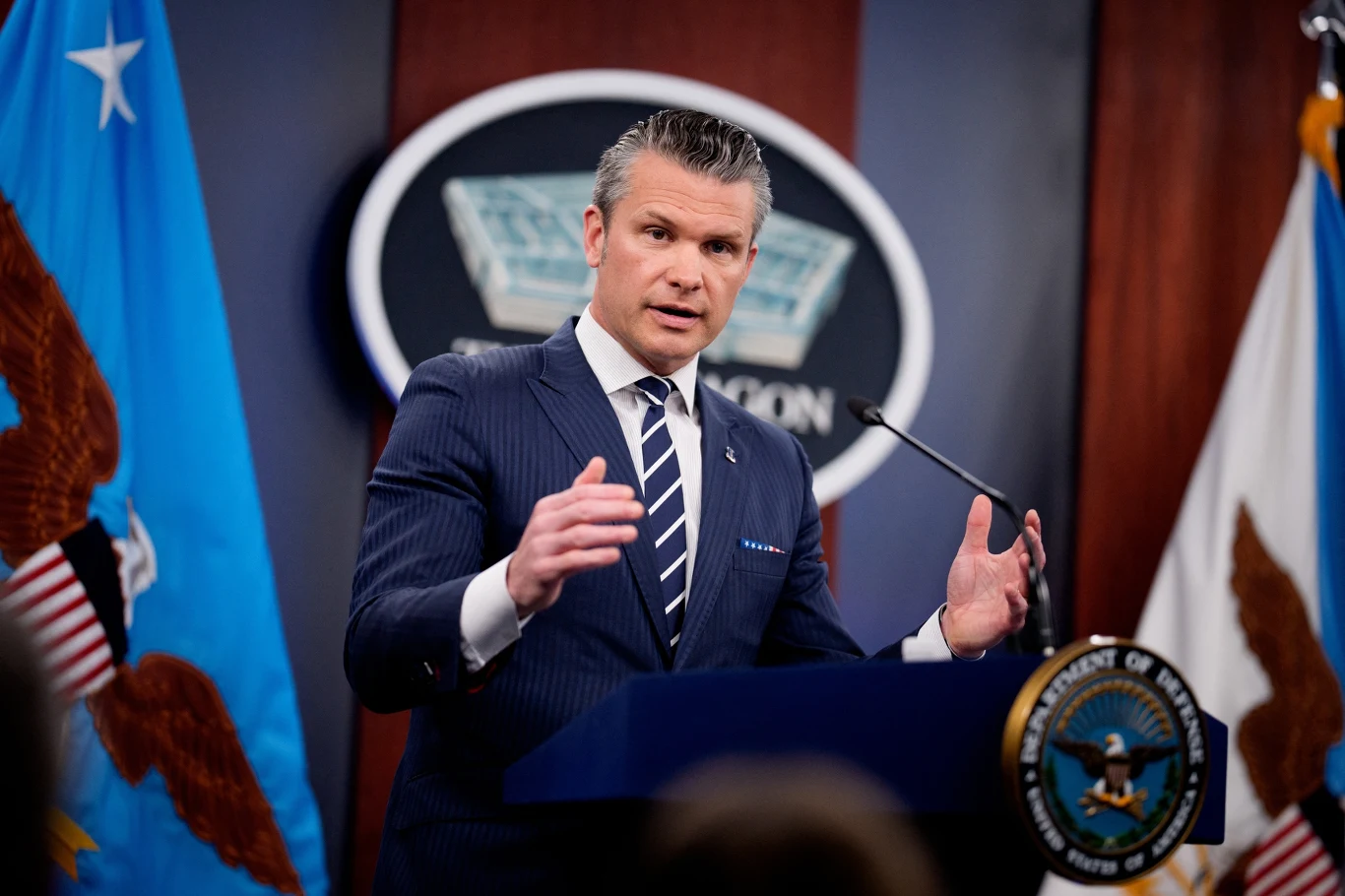 Sekretarz obrony USA Pete Hegseth Mężczyzna w garniturze przemawia na tle logo Pentagonu, stojąc za mównicą z emblematem Departamentu Obrony USA, po bokach widoczne flagi.