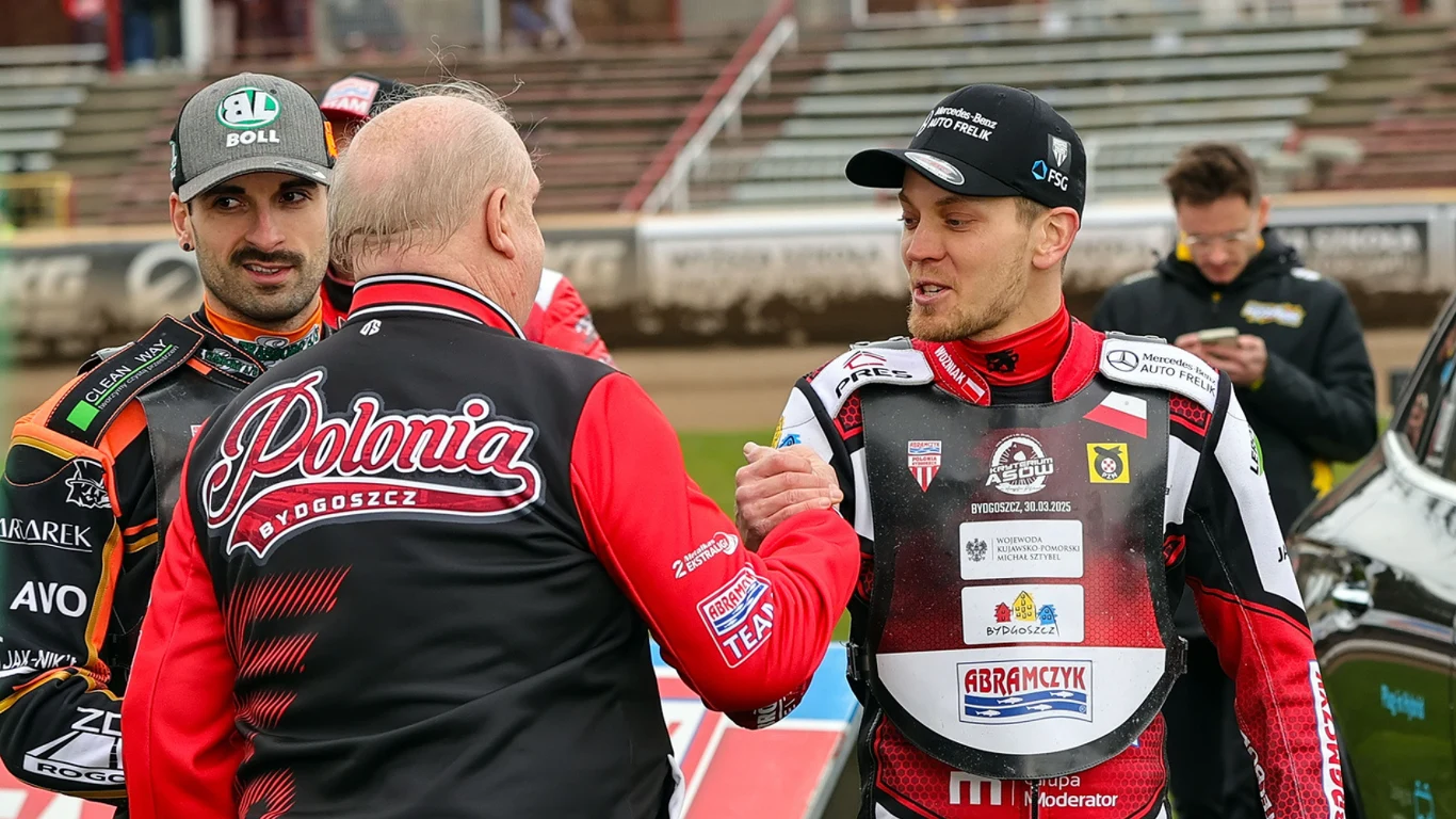 Szymon Woźniak jest liderem Abramczyk Polonii Bydgoszcz Sportowcy w strojach żużlowych ściskający sobie dłonie na torze żużlowym, w tle trybuny stadionu i inni uczestnicy wydarzenia.