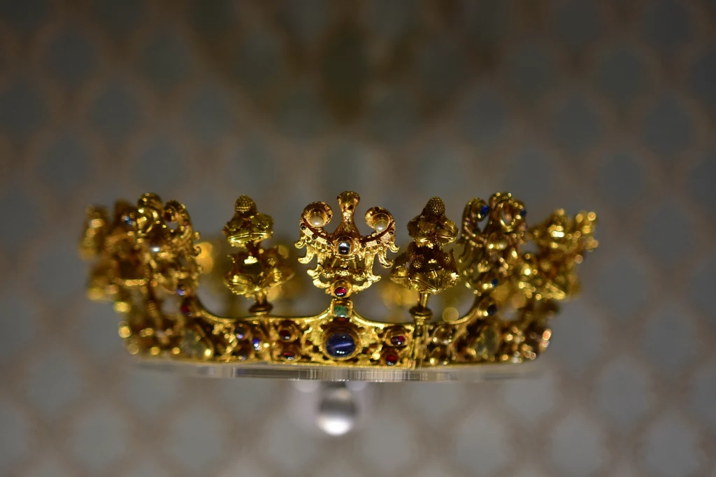 Skarb średzki to jeden z najcenniejszych skarbów odnalezionych w Europie w XX w. Na zdjęciu korona wykonana w XIV w. Złota korona ozdobiona kolorowymi kamieniami szlachetnymi oraz misternie wykonanymi ornamentami, skoncentrowana na centralnym punkcie, lekko rozmyte tło podkreśla blask metalu i klejnotów.