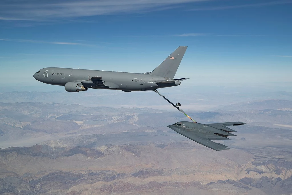 KC-46A Pegasus podczas tankowania z pomocą wysięgnika bombowca B-2 Spirit Samolot wojskowy – tankowiec powietrzny – w trakcie tankowania bombowca strategicznego w powietrzu, na tle rozległego, górzystego krajobrazu pod nimi, niebo czyste z lekkimi smugami chmur.