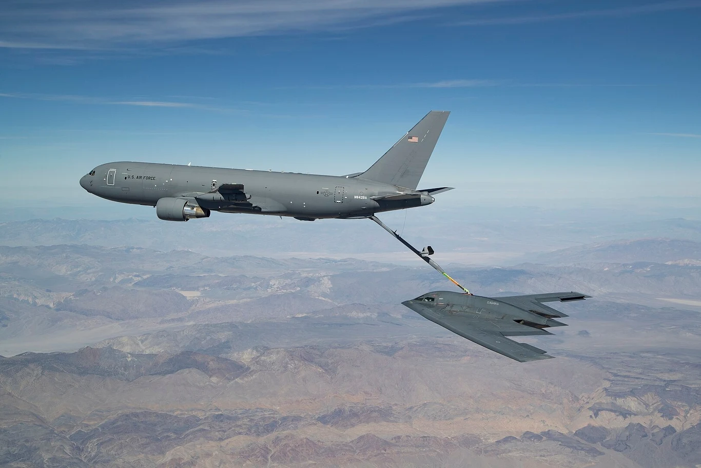 KC-46A Pegasus podczas tankowania z pomocą wysięgnika bombowca B-2 Spirit Samolot wojskowy – tankowiec powietrzny – w trakcie tankowania bombowca strategicznego w powietrzu, na tle rozległego, górzystego krajobrazu pod nimi, niebo czyste z lekkimi smugami chmur.