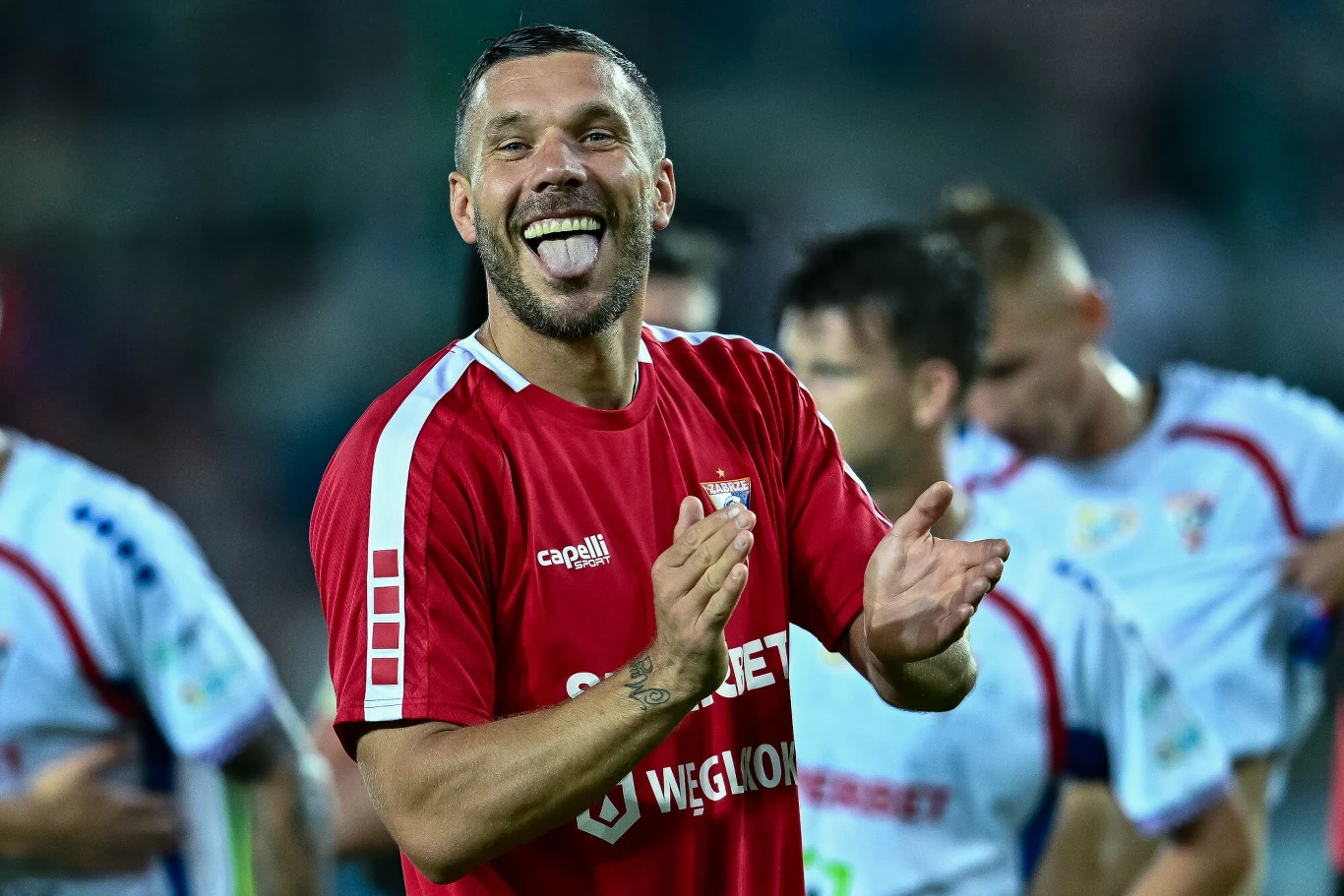 Piłkarz w czerwonej koszulce z uśmiechem na twarzy i wyciągniętym językiem, klaszczący w dłonie, w tle inni piłkarze w strojach sportowych