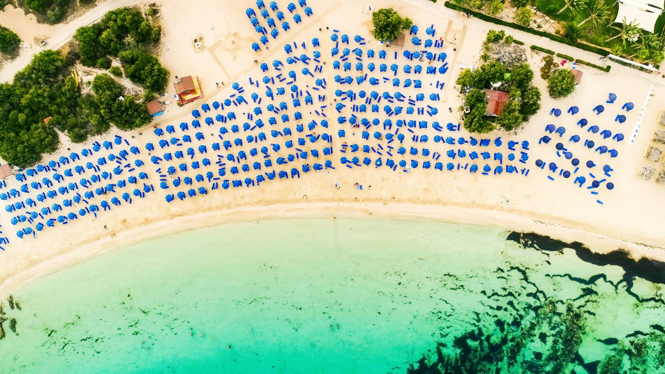 Ayia Napa z lotu ptaka Rzędy niebieskich parasoli ustawionych na piaszczystej plaży przy jasnoturkusowej wodzie, otoczone zielenią i kilkoma budynkami.