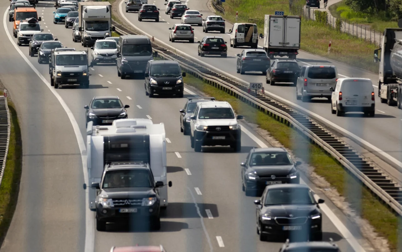 Czy można wyprzedzać z przyczepą na autostradzie? Gęsty ruch samochodów osobowych, dostawczych i ciężarówek na kilkupasmowej drodze, oddzielonej barierami i zielenią, przy bezchmurnej pogodzie.