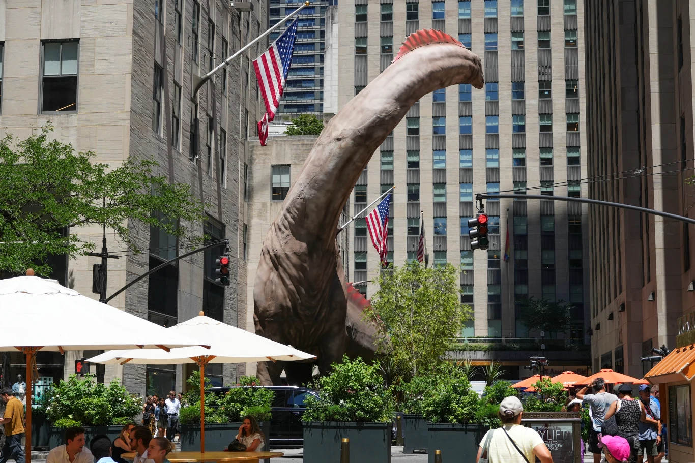 Filmowy tytanozaur z "Jurassic World Rebirth" Ogromna rzeźba dinozaura, najprawdopodobniej diplodoka, wystaje spośród miejskich zabudowań i drzew, otoczona flagami USA oraz przechodniami spacerującymi w pobliżu kawiarnianych parasoli.