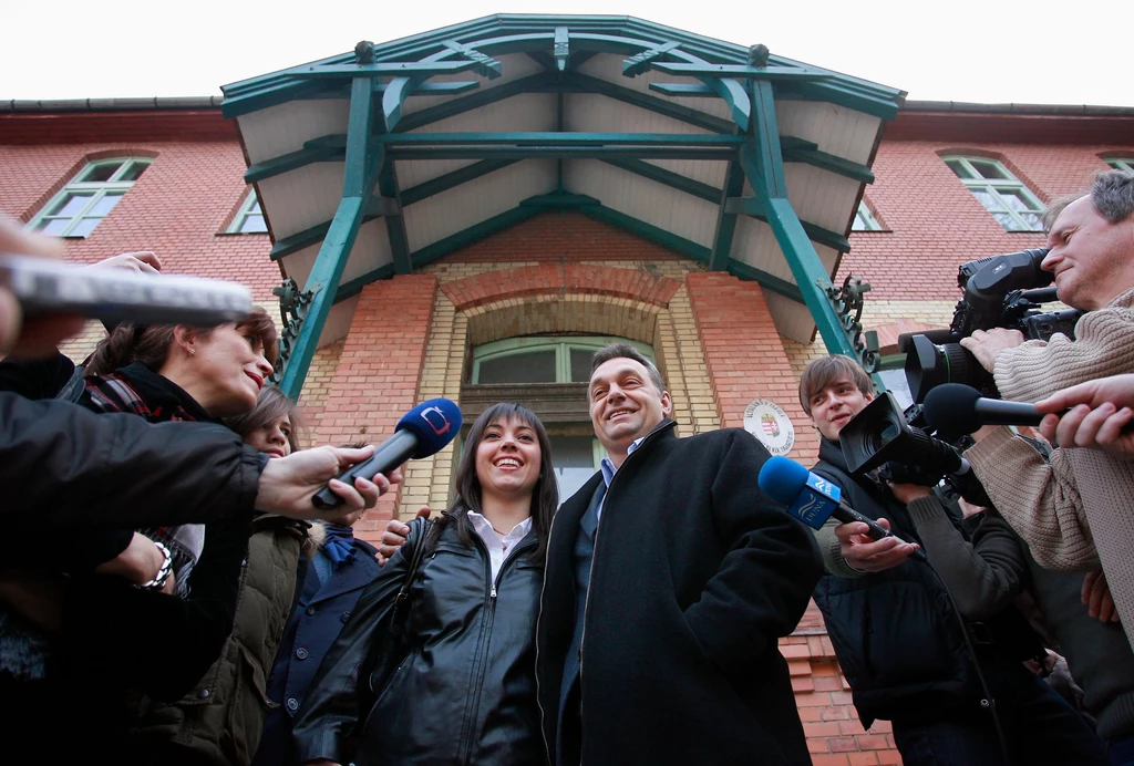Para uśmiechniętych osób otoczona przez grupę dziennikarzy trzymających mikrofony i kamery, na tle czerwonego budynku z widocznym wejściem i charakterystycznym daszkiem.