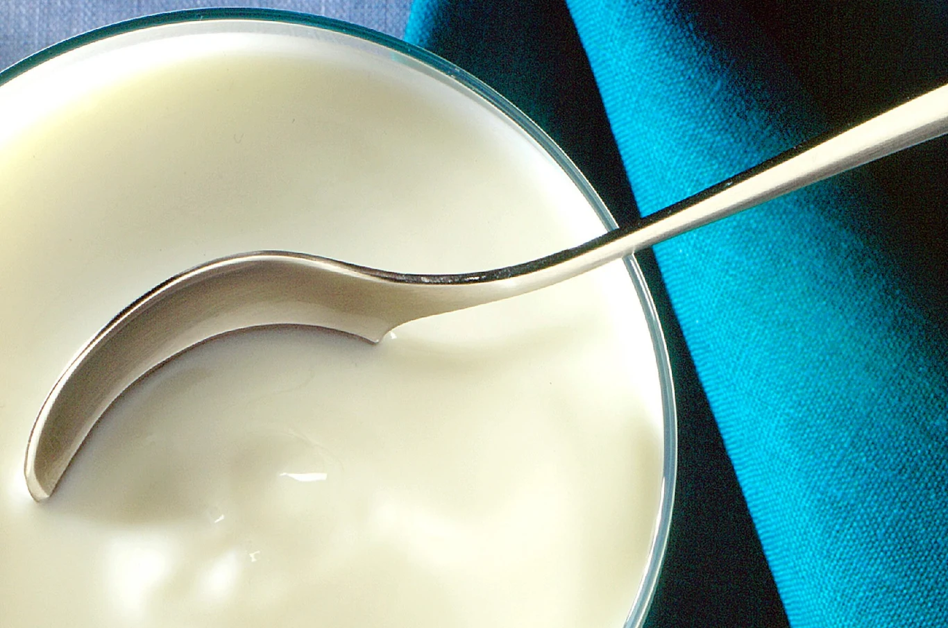 Kefir warto jeść jak najczęściej. Należy jednak dobrze przemyśleć dodatki Metalowa łyżka zanurzona w gładkim, kremowym jogurcie, w tle widoczny niebiesko-turkusowy materiał.