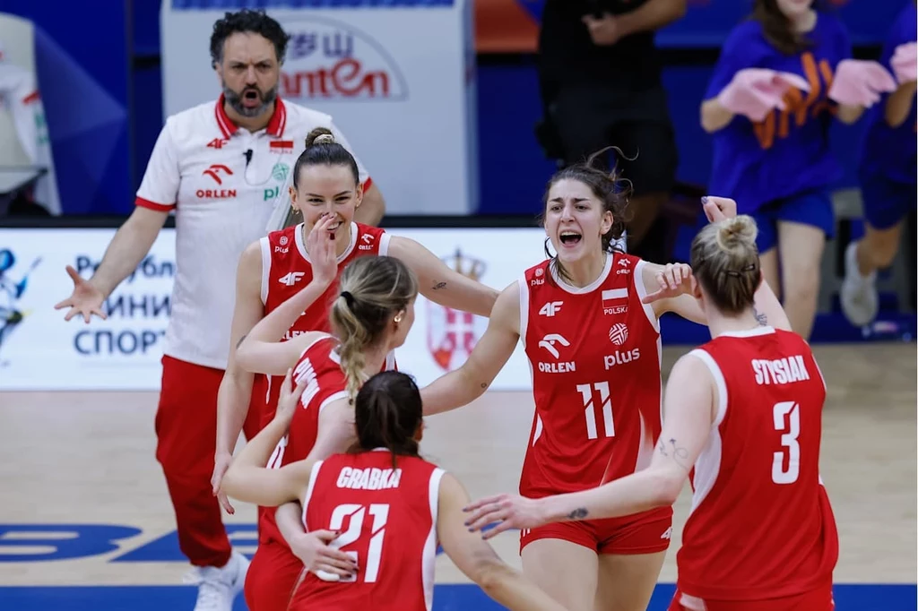Siatkarki reprezentacji Polski Grupa siatkarek w czerwonych strojach z numerami świętuje razem na boisku, wyrażając radość i entuzjazm, za nimi trener również reaguje emocjonalnie.