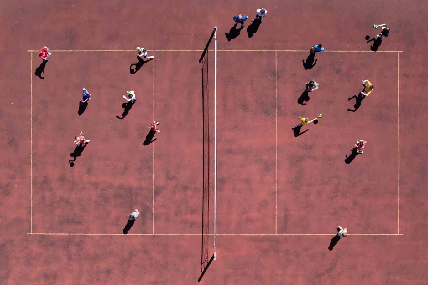 Grupa ludzi na boisku do siatkówki, ujęcie z lotu ptaka, wyraźnie widoczne linie boiska, siatka umieszczona pośrodku oraz cienie osób grających.