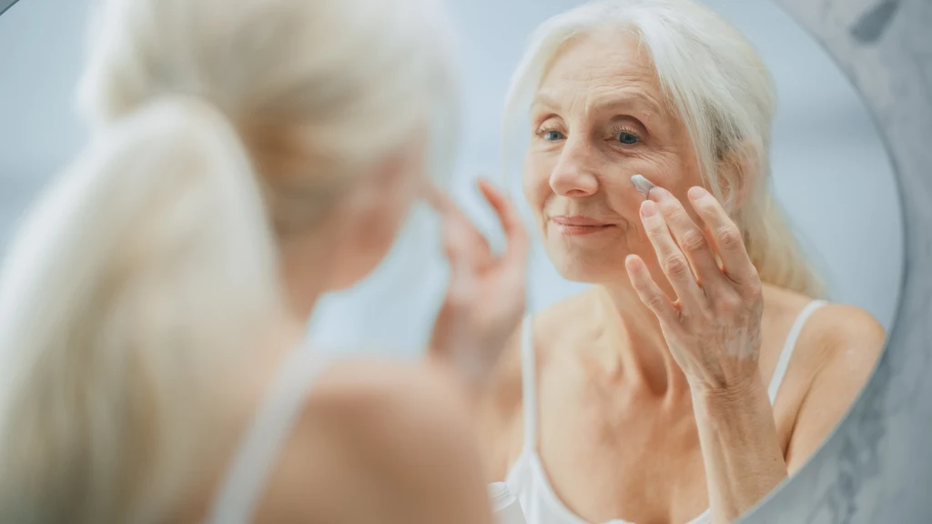 Choć retinol pomaga zachować cerę w doskonałym stanie, to może źle reagować na ślubie Starsza kobieta nakłada krem na twarz, spoglądając w lustro, uśmiechnięta, w jasnym, naturalnym otoczeniu, w białej koszulce na ramiączkach.