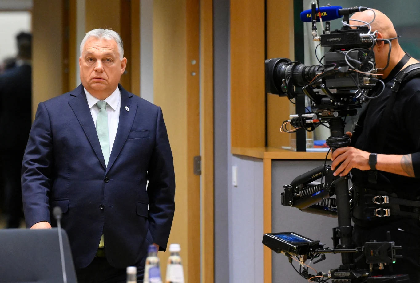 Viktor Orban Mężczyzna w garniturze ze spokojnym wyrazem twarzy stoi w pomieszczeniu konferencyjnym, obok niego znajduje się operator z dużą kamerą filmową i sprzętem nagrywającym.
