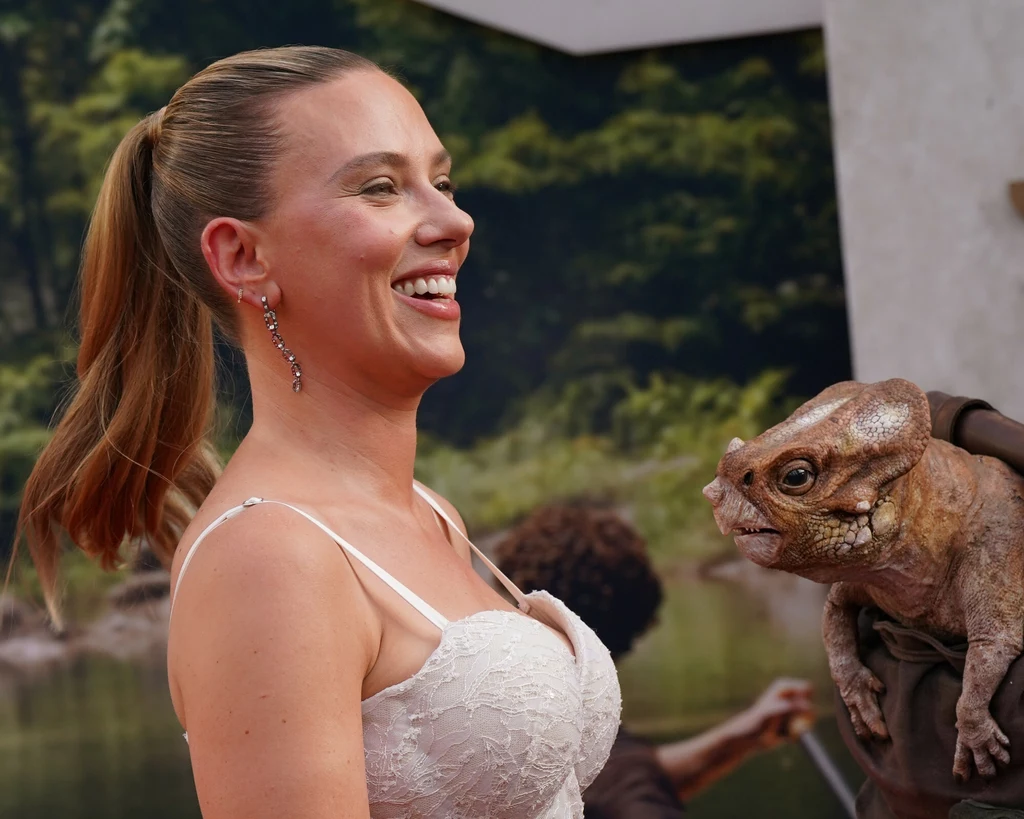 Scarlett Johansson i Aquilops z filmu "Jurassic World. Rebirth" Kobieta w jasnej sukience na tle zieleni uśmiecha się szeroko, obok niej znajduje się realistyczna figurka lub lalka prehistorycznego gada.