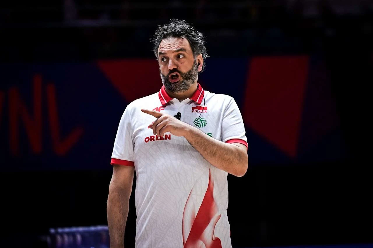 Trener w białej koszulce sportowej Orlen gestykuluje podczas wydarzenia sportowego, wyrazisty wyraz twarzy świadczący o intensywnej komunikacji lub przekazywaniu instrukcji, ciemne tło z nieostrymi kolorowymi elementami.