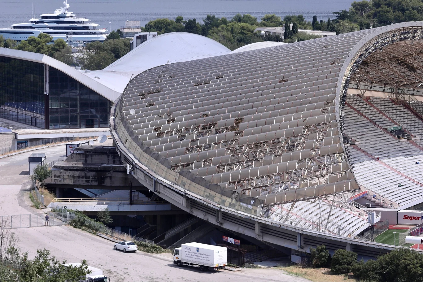 Uszkodzony dach stadionu z rozległymi zniszczeniami pokrycia, puste trybuny oraz widoczne obiekty pomocnicze w otoczeniu, w tle duży jacht na wodzie oraz zabudowania i zieleń.