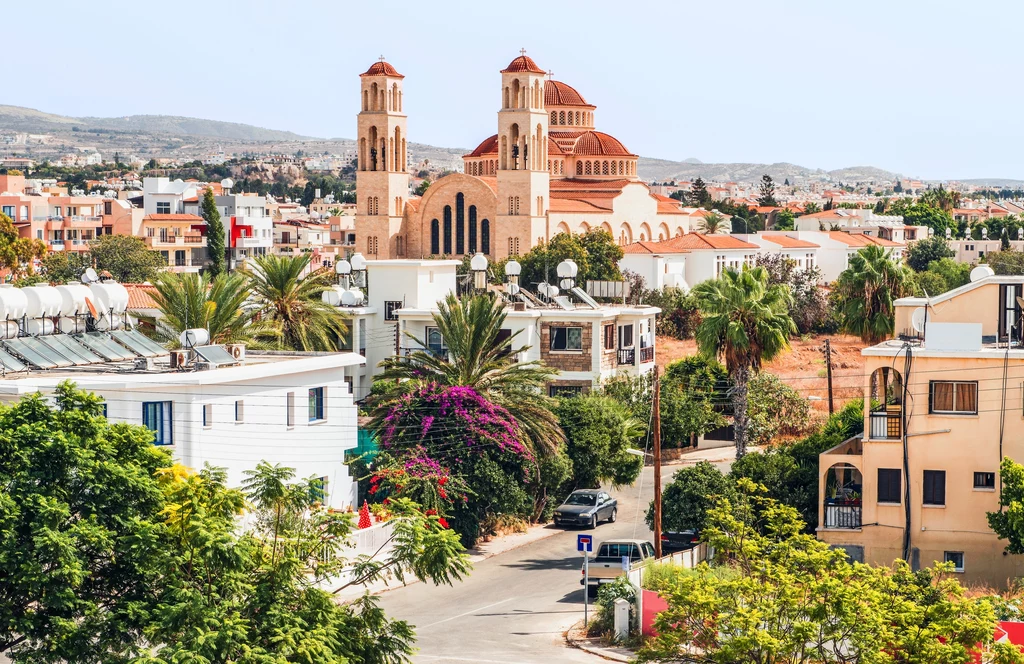 Pafos Duży kościół z dwoma wieżami górujący nad zielonym i słonecznym miastem, otoczony nowoczesnymi budynkami oraz palmami i krzewami.