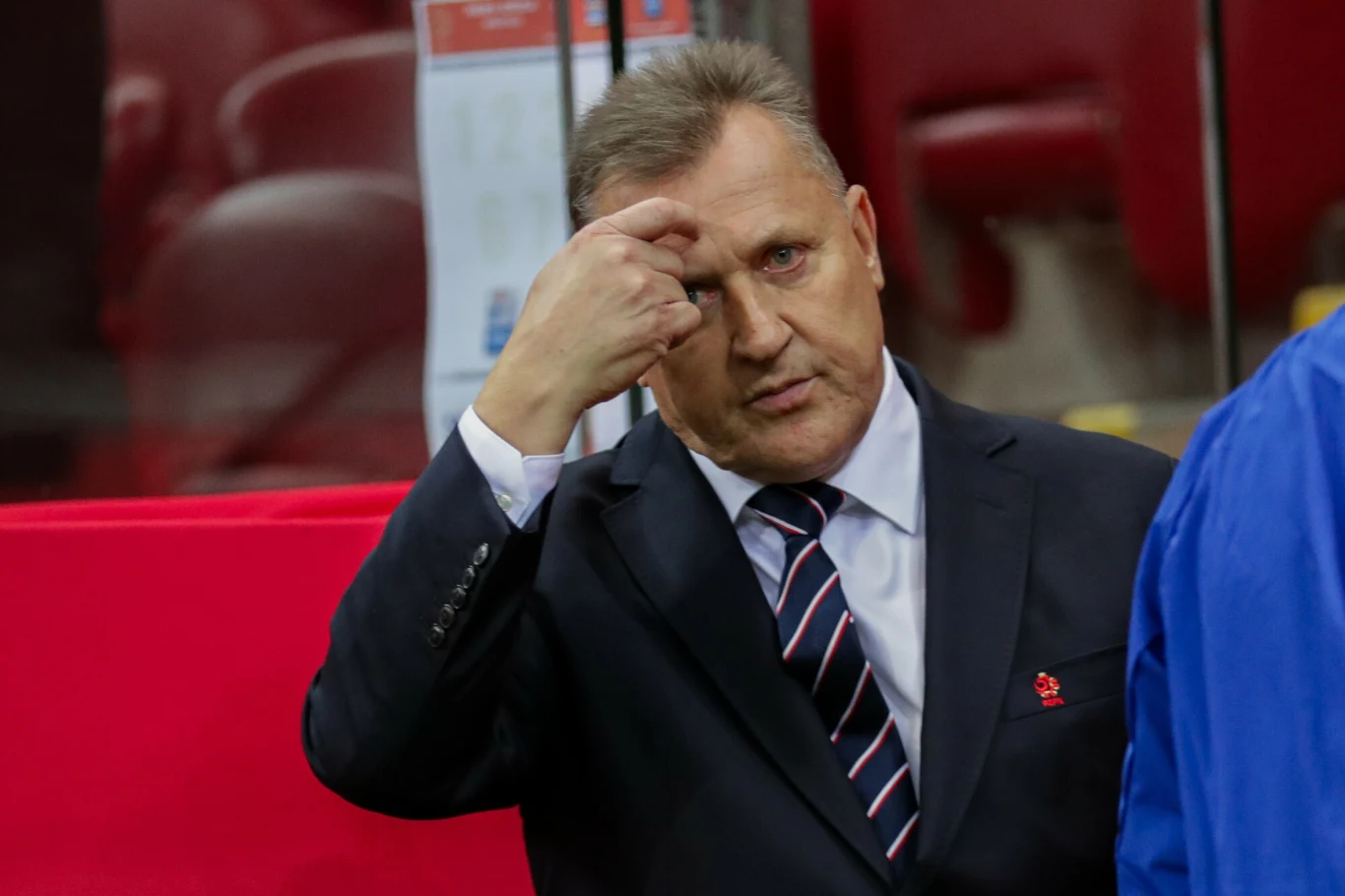 Mężczyzna w garniturze i krawacie wskazuje palcem na czoło, stojąc na tle czerwonych siedzeń stadionowych.