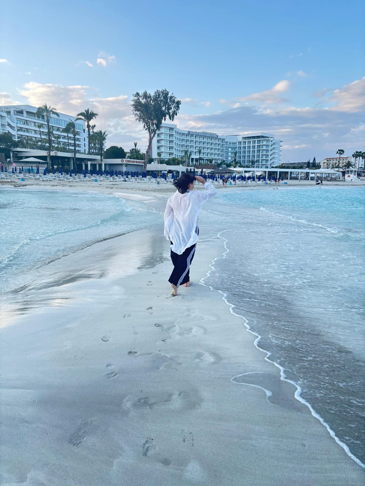 Nissi Osoba idąca boso po piaszczystej plaży w kierunku luksusowego hotelu, ubrana w białą koszulę i ciemne spodnie. W tle widać budynki hotelowe otoczone palmami i ludzi spędzających czas przy brzegu morza.