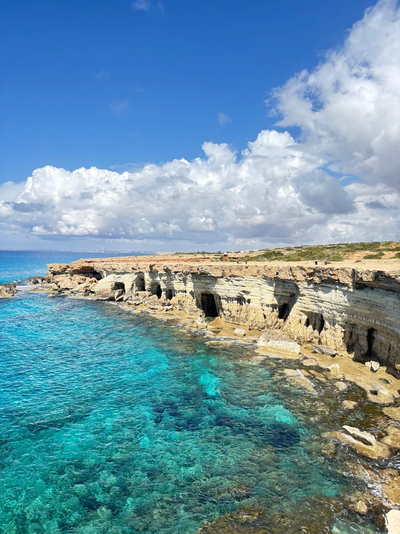 Cape Greco Malowniczy klif z jasnych skał opadających do turkusowego morza pod błękitnym niebem z dynamicznymi chmurami; na szczycie klifu widoczny wąski pas roślinności.